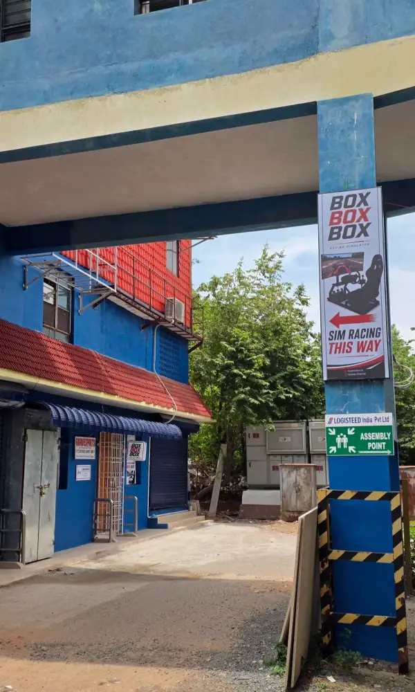 The BOXBOXBOX banner on the pillar at the entrance to the Readymade Garments Complex in Guindy, Chennai, with a red arrow reading SIM RACING THIS WAY pointing down the lane to the lounge.